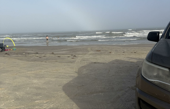 Sandy beach at Padre Island National Seashore with the corner of an automobile showing and a child wading in large waves.