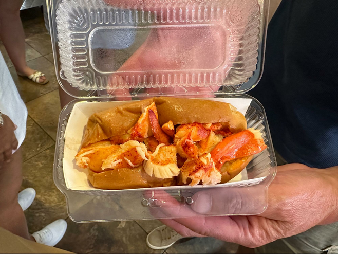 Hands hold an open to-go container with a lobster roll inside.